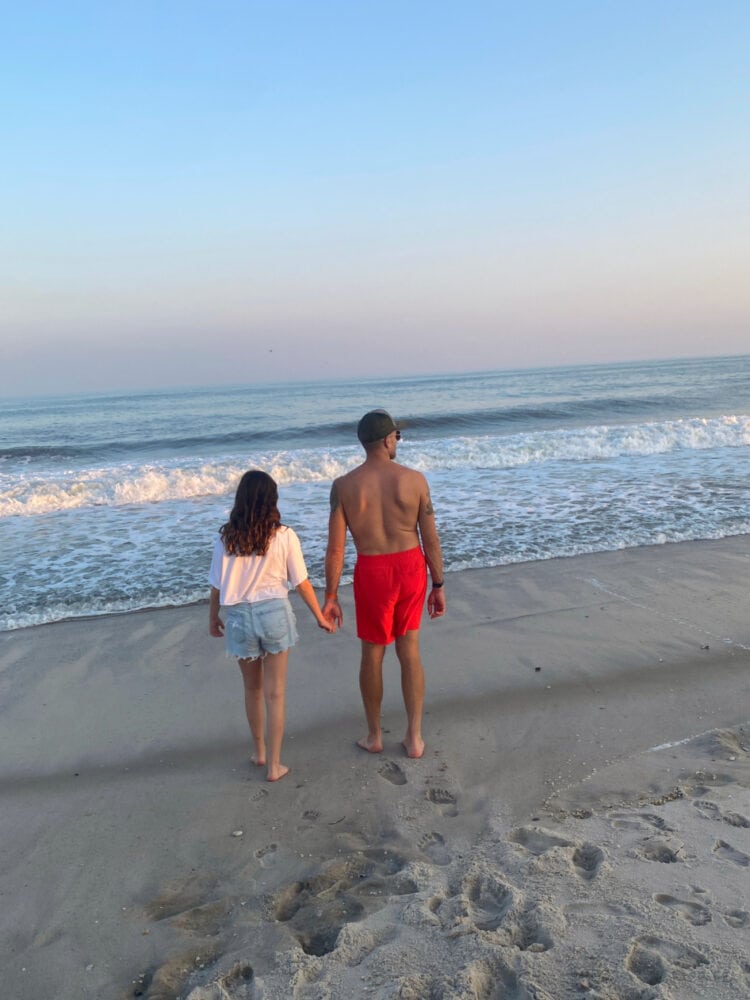 A man and his tween daughter holding hands, standing on a beach, looking at the sunset